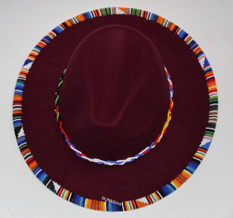 May include: A burgundy fedora-style hat with a wide brim. The brim is adorned with a colorful beaded trim. A smaller beaded band encircles the crown. The hat has the text "MJN&Sons" on the brim.