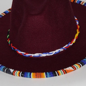 May include: A burgundy fedora hat with a wide brim, adorned with colorful beaded trim around the crown and brim. The hat features a braided beaded band in red, white, and blue. The hat's design is a fusion of style and cultural elements.