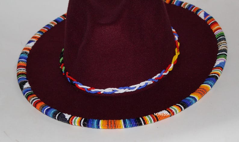 May include: A burgundy fedora hat with a wide brim, adorned with colorful beaded trim around the crown and brim. The hat features a braided beaded band in red, white, and blue. The hat's design is a fusion of style and cultural elements.