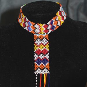 May include: A vibrant beaded necklace with a geometric design. The necklace has a collar shape with a central pendant. The beads are in a variety of colours, including red, yellow, blue, orange, and white. The necklace is displayed on a black mannequin.