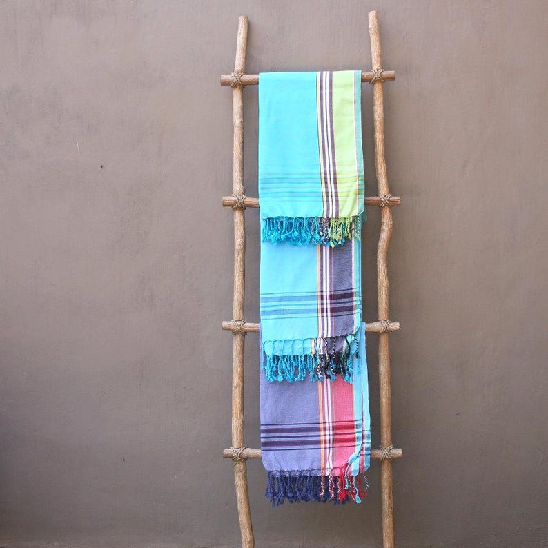 Op de afbeelding: Drie kleurrijke Turkse handdoeken gedrapeerd over een houten ladder. De handdoeken zijn turquoise, blauw en paars, met gestreepte patronen en gefranjerde randen. De ladder is gemaakt van natuurlijk hout en leunt tegen een neutraal gekleurde muur.