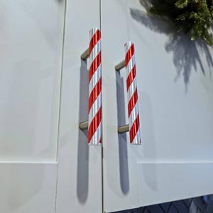 Peut inclure: Deux portes d'armoires blanches avec des poignées à rayures de canne en sucre. Les poignées sont rouges et blanches, cylindriques et montées sur du matériel argenté. Les portes d'armoires sont blanches et ont un design simple et moderne. Une couronne de Noël est visible en arrière-plan.