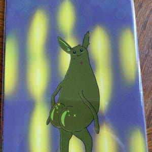 Puede incluir: Una lámina de arte rectangular que muestra una criatura verde de dibujos animados con orejas grandes y una cara sonriente. La criatura se representa desde atrás, mostrando su parte trasera redondeada con reflejos brillantes. El fondo es morado con haces de luz amarillos.
