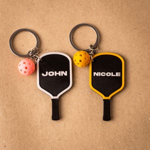 Könnte beinhalten: Zwei personalisierte Pickleball-Schlüsselanhänger. Ein Schläger ist schwarz mit weißem Rand und dem Namen "JOHN" in Weiß. Der andere ist schwarz mit gelbem Rand und dem Namen "NICOLE" in Weiß. Jeder Schlüsselanhänger hat einen kleinen, farbigen Ball.