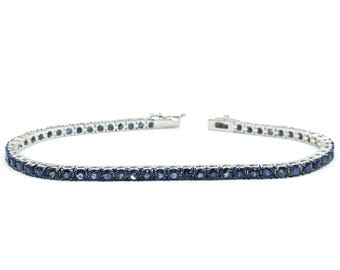 May include: A silver tennis bracelet with a row of dark blue gemstones. The bracelet is set against a white background, showcasing the jewelry's design and color. The clasp is open, showing the bracelet's construction.