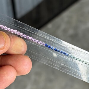 Puede incluir: Una cadena de pequeñas piedras preciosas de colores en un paquete de plástico transparente. Las piedras preciosas cambian de color de rosa a azul a verde. Las piedras preciosas son pequeñas y redondas, probablemente para la fabricación de joyas.