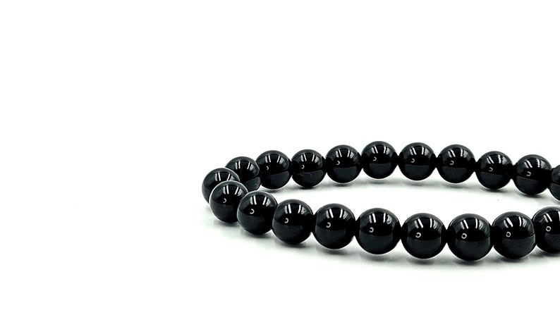 May include: A bracelet composed of glossy, black, spherical beads. The beads are strung together, forming a circular shape. The bracelet is set against a plain white background, highlighting the dark, reflective surfaces of the beads. Jewelry.