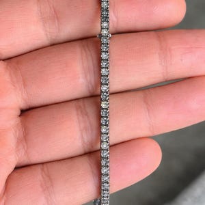 Puede incluir: Una delicada pulsera de plata con una fila de pequeñas piedras preciosas brillantes. La pulsera se sostiene en una mano, mostrando su diseño delgado y el brillo de las piedras. Las piedras preciosas miden aproximadamente 0,25 cm de diámetro.