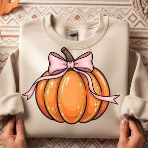 May include: Cream-colored sweatshirt featuring a large, illustrated orange pumpkin with a pink bow and glitter accents. The pumpkin has a brown stem and is centered on the front of the long-sleeved top. A fall-themed design.