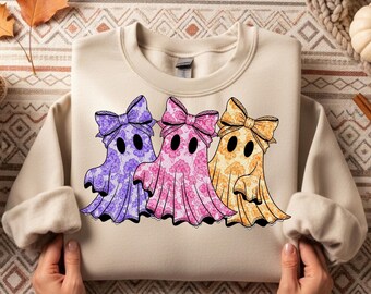 May include: A beige sweatshirt featuring three ghost illustrations with floral patterns in purple, pink, and orange. Each ghost has a matching bow and black eyes. The sweatshirt is displayed on a patterned surface.