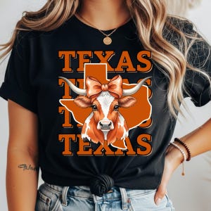 Könnte beinhalten: Schwarzes T-Shirt mit einer Grafik einer Longhorn-Kuh mit Schleife, vor einem orangefarbenen Texas-Umriss. Das Wort "TEXAS" ist dreimal in orangefarbenen Buchstaben über und unter der Kuh-Illustration gestapelt.