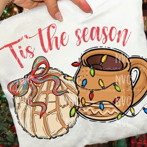May include: White t-shirt with the text "Tis the season" in red script. The design features a brown mug with colourful Christmas lights and a pastry with a red, blue, and green bow.