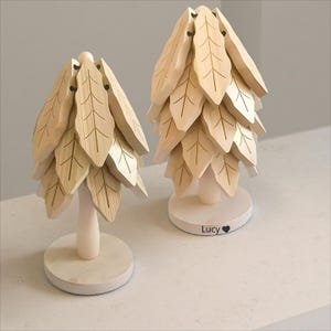 Könnte beinhalten: Zwei dekorative Bäume aus Holz in Blattform. Die Bäume sind aus hellfarbenem Holz gefertigt und haben ein geschichtetes Design, das Blättern ähnelt. Jeder Baum steht auf einem weißen, kreisförmigen Sockel. Auf dem Sockel eines Baumes steht der Name "Lucy".