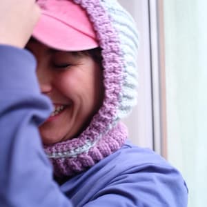 Könnte beinhalten: Eine rosa Baseballkappe wird unter einer gehäkelten Kapuze in Lila- und Hellblautönen getragen. Die Person trägt eine blaue Jacke. Die Kapuze liegt um Hals und Gesicht.