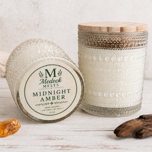 May include: Two glass candle jars with textured beaded patterns and wooden lids. One jar stands upright, while the other lies on its side, displaying a label that reads M Medeck Melts Midnight Amber, hand-poured, natural soy wax, 10 oz.