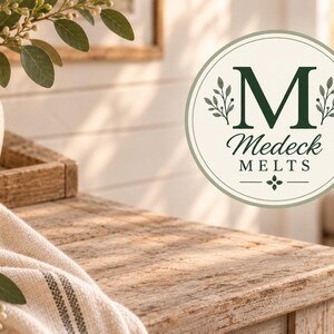 May include: A circular logo featuring a large dark green letter M, flanked by small leaf sprigs, with the text Medeck MELTS below it. The logo is set against a light cream background with a thin green border, presented in a bright, sunlit room.