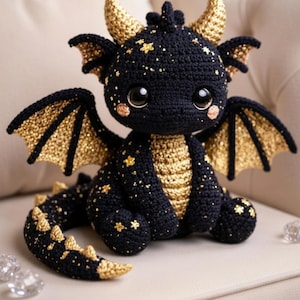 Könnte beinhalten: Ein handgehäkelter Drache in Schwarz und Gold. Der Drache hat große, runde Augen, kleine Flügel und einen langen Schwanz. Goldene Sterne sind über den Körper des Drachen verstreut. Der Drache sitzt auf einer cremefarbenen Oberfläche.