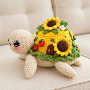 Könnte beinhalten: Ein gehäkeltes Schildkröten-Plüschtier mit einem gelben Panzer, der mit Sonnenblumen, Erdbeeren und einer kleinen braunen Tür verziert ist. Die Schildkröte hat einen cremefarbenen Körper, Beine und Kopf mit schwarzen Augen und rosa Wangen. Das Plüschtier liegt auf einem cremefarbenen Sofa.