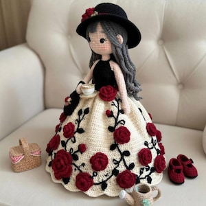 Könnte beinhalten: Gehäkelte Puppe mit schwarzem Hut mit roten Rosenakzenten, schwarzem Oberteil und cremefarbenem Kleid, verziert mit roten Rosen und schwarzen Ranken. Zubehör: kleine Handtasche, Teetasse, Korb, Gießkanne und rote Schuhe.