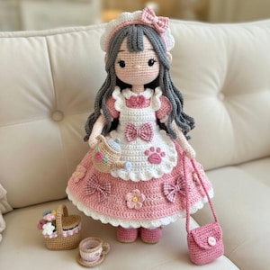 Könnte beinhalten: Handgefertigte Häkelpuppe in einem rosa Kleid mit weißen Akzenten, passendem Stirnband und einer kleinen rosa Handtasche. Die Puppe hat graues Haar und hält einen Korb und ein Teeservice.