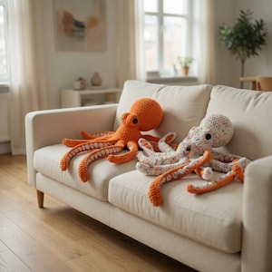 Könnte beinhalten: Zwei gehäkelte Oktopus-Plüschtiere in Orange und bunten Mustern liegen auf einem cremefarbenen Sofa. Der orangefarbene Oktopus hat eine einfarbige Farbe, während der andere eine Mischung aus Pastellfarben aufweist. Beide haben lange, gekräuselte Tentakel.