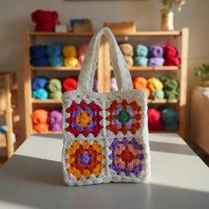 Könnte beinhalten: Eine handgefertigte, cremefarbene Häkeltasche mit vier bunten Granny Squares. Die Quadrate zeigen leuchtende Farbtöne von Orange, Lila, Rot und Grün. Die Tasche hat zwei stabile Griffe und wird auf einer hellen Oberfläche gezeigt.