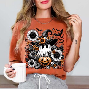 May include: Orange t-shirt featuring a Halloween-themed graphic. The design includes a ghost wearing a witch hat, surrounded by sunflowers, pumpkins, and bats. The shirt is a solid orange color.