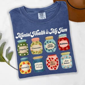 May include: A blue t-shirt with the phrase "Mental Health is My Jam" in white script. The shirt features illustrations of jars labeled with terms like "Empathy," "Communication," and "Mindfulness." The shirt is a Comfort Colors brand.