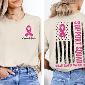 May include: Beige t-shirt with a pink ribbon graphic and the text "#TeamOlivia" on the front. The back of the shirt features a pink ribbon, stars, and stripes design with the words "SUPPORT SQUAD" and "BREAST CANCER AWARENESS".