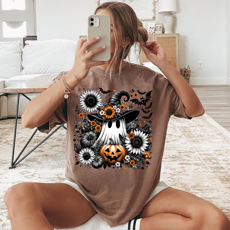May include: A brown t-shirt featuring a Halloween-themed graphic. The design includes a ghost wearing a witch hat, surrounded by sunflowers, pumpkins, and bats. The artwork is in shades of black, white, orange, and yellow.