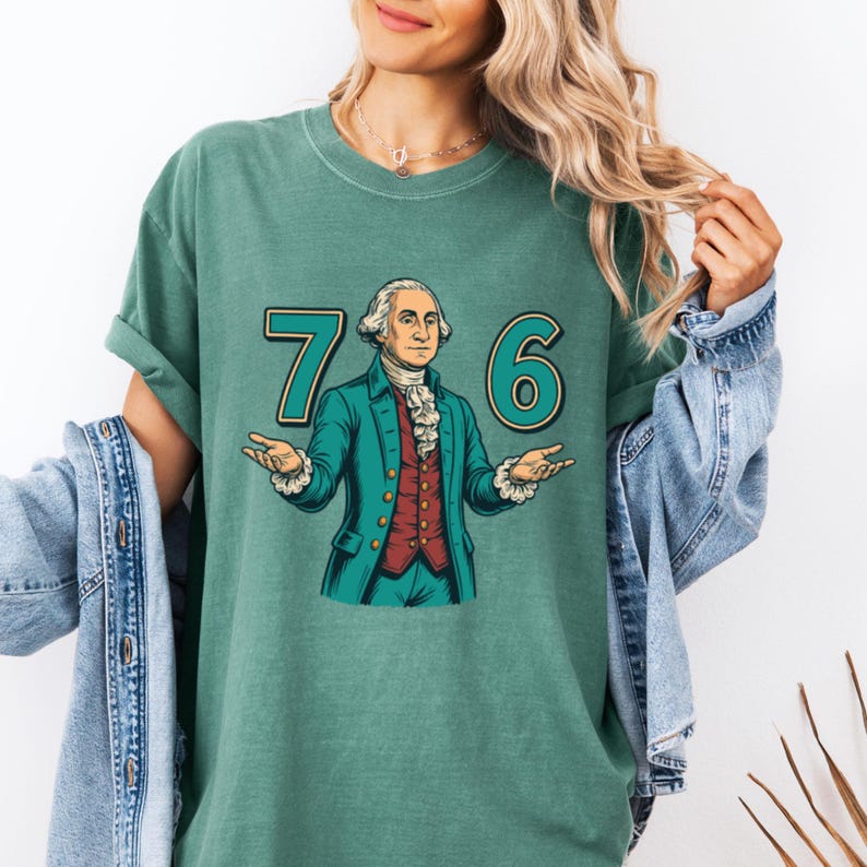 Puede incluir: Una camiseta de color verde azulado con un gr&aacute;fico de George Washington y los n&uacute;meros "76" en verde azulado. Washington est&aacute; representado con un abrigo verde azulado, un chaleco rojo y una camisa blanca con volantes. La camiseta es de estilo casual.