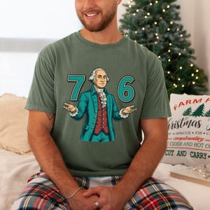 Puede incluir: Camiseta verde con un gr&aacute;fico de George Washington y los n&uacute;meros "76" en color turquesa. Washington lleva una chaqueta turquesa, un chaleco rojo y una corbata blanca. La camiseta es de color verde liso.