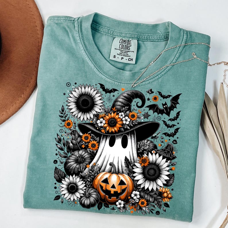 May include: A teal Comfort Colors t-shirt featuring a Halloween-themed graphic. The design includes a ghost wearing a witch hat, surrounded by sunflowers, pumpkins, and bats. The shirt has a crew neck and short sleeves. The tag reads "S - P - CH".