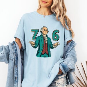 Puede incluir: Camiseta azul claro con un gr&aacute;fico de George Washington y los n&uacute;meros "7" y "6" en verde azulado. Washington lleva un abrigo verde azulado, un chaleco rojo y una camisa blanca con volantes. La camiseta es de estilo casual.