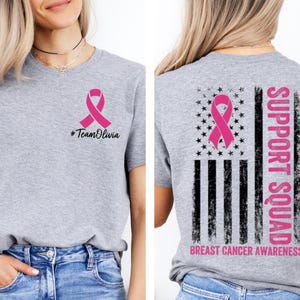 May include: A heather gray t-shirt with a pink ribbon graphic, the text "#TeamOlivia" on the front, and a distressed American flag design with a pink ribbon and the words "Support Squad" and "Breast Cancer Awareness" on the back.