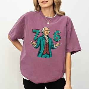 Puede incluir: Camiseta de color malva con un gr&aacute;fico de George Washington y los n&uacute;meros "76" en verde azulado. Washington lleva una chaqueta verde azulado y un chaleco rojo, con los brazos extendidos. La camiseta es de estilo casual y oversize.