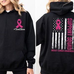 May include: Black hoodie with a pink ribbon and the text "#TeamOlivia" on the front. The back features a pink ribbon over a distressed American flag design with "SUPPORT SQUAD" and "BREAST CANCER AWARENESS" text.
