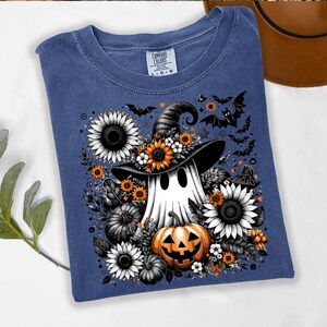 May include: A blue t-shirt featuring a Halloween-themed graphic. The design includes a ghost wearing a witch hat, surrounded by sunflowers, pumpkins, and bats. The shirt has a crew neck and the Comfort Colors brand label.