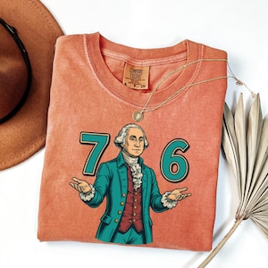 Puede incluir: Una camiseta Comfort Colors de color coral con un gr&aacute;fico de George Washington y los n&uacute;meros "76" en verde azulado. La ilustraci&oacute;n muestra a Washington con un abrigo verde azulado y un chaleco rojo. Tambi&eacute;n se ve un collar dorado y un sombrero marr&oacute;n.