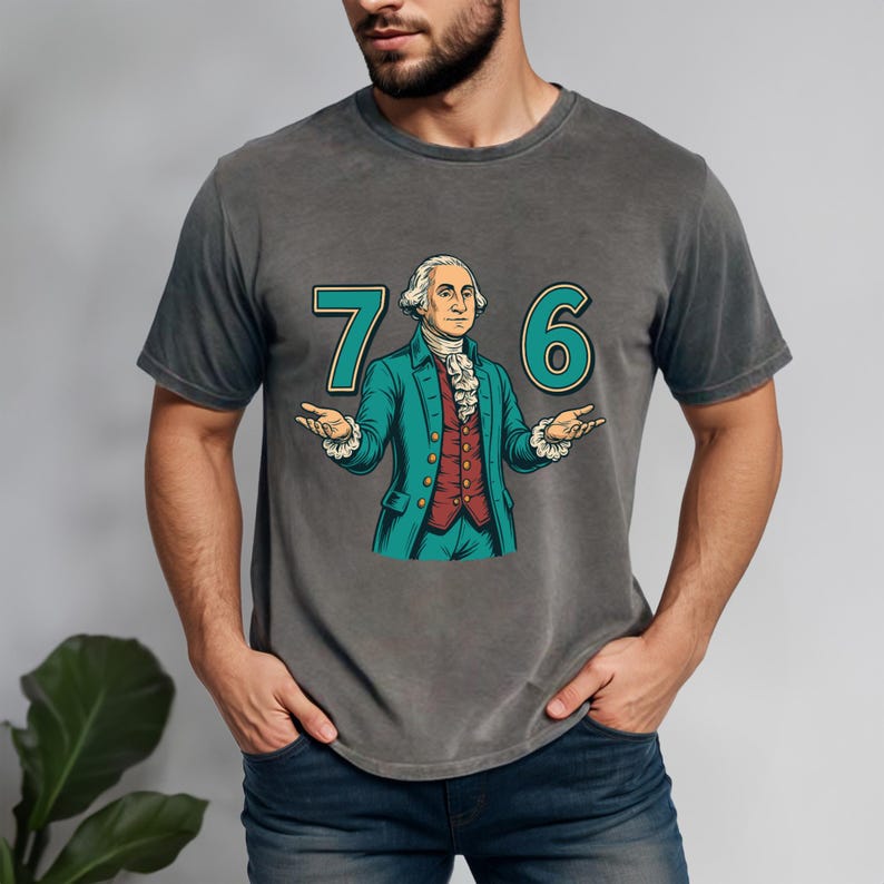Puede incluir: Camiseta gris con una ilustraci&oacute;n de dibujos animados de George Washington y los n&uacute;meros "76" en verde azulado. Washington lleva un abrigo verde azulado y un chaleco rojo, con los brazos extendidos. La camiseta es de estilo casual.