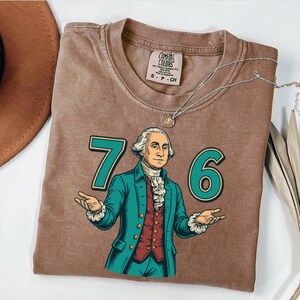 Puede incluir: Camiseta marr&oacute;n con un gr&aacute;fico de George Washington y los n&uacute;meros "76" en verde azulado. La camiseta tiene un aspecto vintage, con una etiqueta Comfort Colors. Tambi&eacute;n se ve un collar plateado con un colgante.