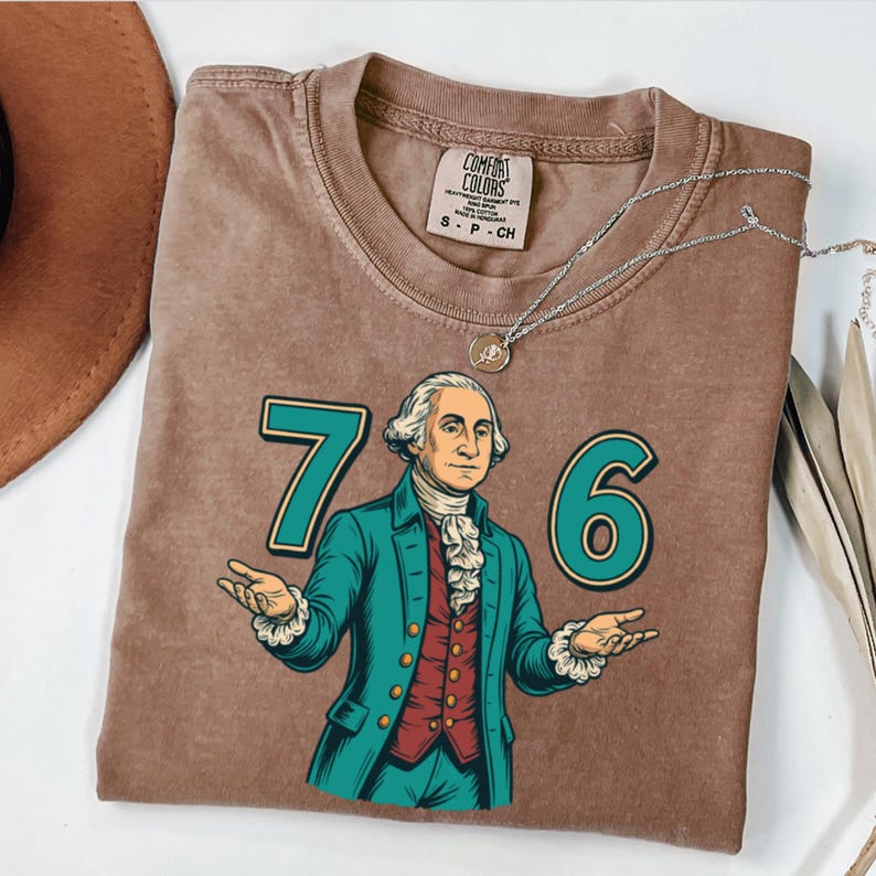 Puede incluir: Camiseta marr&oacute;n con un gr&aacute;fico de George Washington y los n&uacute;meros "76" en verde azulado. La camiseta tiene un aspecto vintage, con una etiqueta Comfort Colors. Tambi&eacute;n se ve un collar plateado con un colgante.