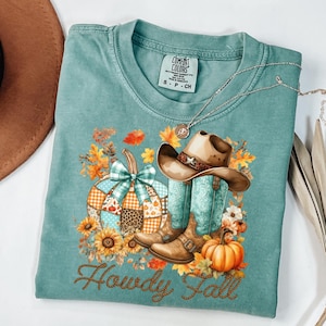 May include: A teal Comfort Colors t-shirt featuring a fall-themed graphic. The design includes a pumpkin, cowboy boots, a hat, and the words "Howdy Fall". The shirt also has a small tag that says "Comfort Colors". A silver necklace is draped across the shirt.