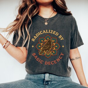 May include: Dark gray t-shirt with the text "RADICALIZED BY BASIC DECENCY" in a circular design. The design features a brown raven inside a teal, orange, and yellow floral mandala. The shirt is a casual, comfortable style.