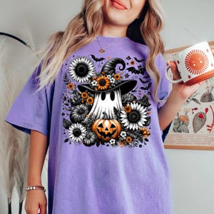 May include: A lilac-colored t-shirt featuring a Halloween-themed graphic. The design includes a ghost wearing a witch hat, surrounded by sunflowers, pumpkins, and bats. The ghost is holding a jack-o'-lantern. The shirt has a relaxed fit.