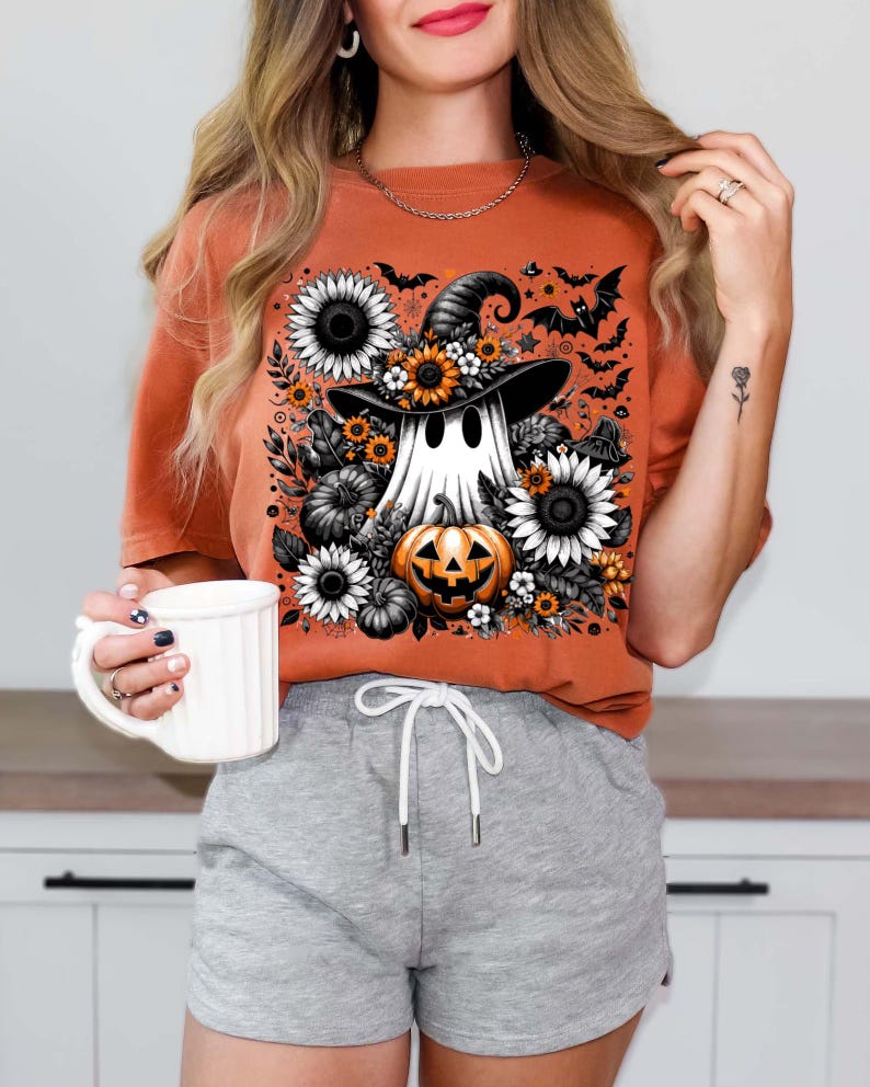 May include: Orange t-shirt featuring a Halloween-themed graphic. The design includes a ghost wearing a witch hat, surrounded by sunflowers, pumpkins, and bats. The shirt is a solid orange color.