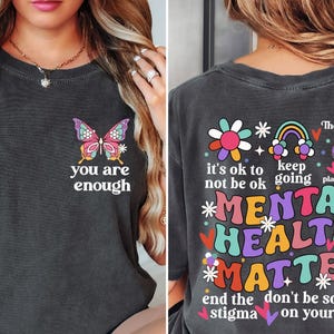 May include: A charcoal gray t-shirt featuring a butterfly and the text "you are enough" on the front. The back displays colorful graphics of a flower, rainbow, and earth with the text "it's ok to not be ok", "keep going", and "MENTAL HEALTH MATTER".