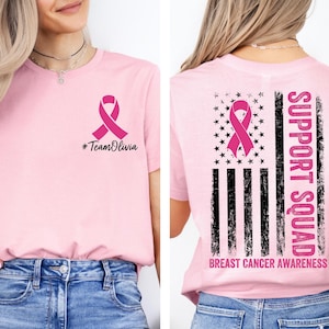 May include: Pink t-shirt with a pink ribbon graphic and the text "#TeamOlivia" on the front. The back features a pink ribbon, stars, stripes, and the words "Support Squad" and "Breast Cancer Awareness".