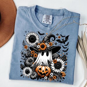 May include: A light blue Comfort Colors t-shirt featuring a Halloween-themed graphic. The design includes a ghost wearing a witch hat, surrounded by sunflowers, pumpkins, and bats. The shirt has a crew neck and short sleeves.