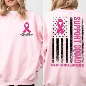 May include: Light pink sweatshirt with a pink ribbon and the text "#TeamOlivia" on the front. The back features a pink ribbon over a black and white flag design with the words "SUPPORT SQUAD" and "BREAST CANCER AWARENESS".